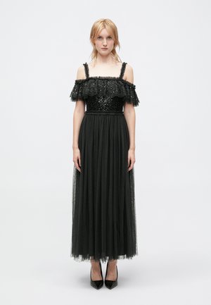 Robe noire avec des manches à volants dénudées, un corsage en sequins et une longue jupe en tulle. Associée à des talons aiguilles noirs.