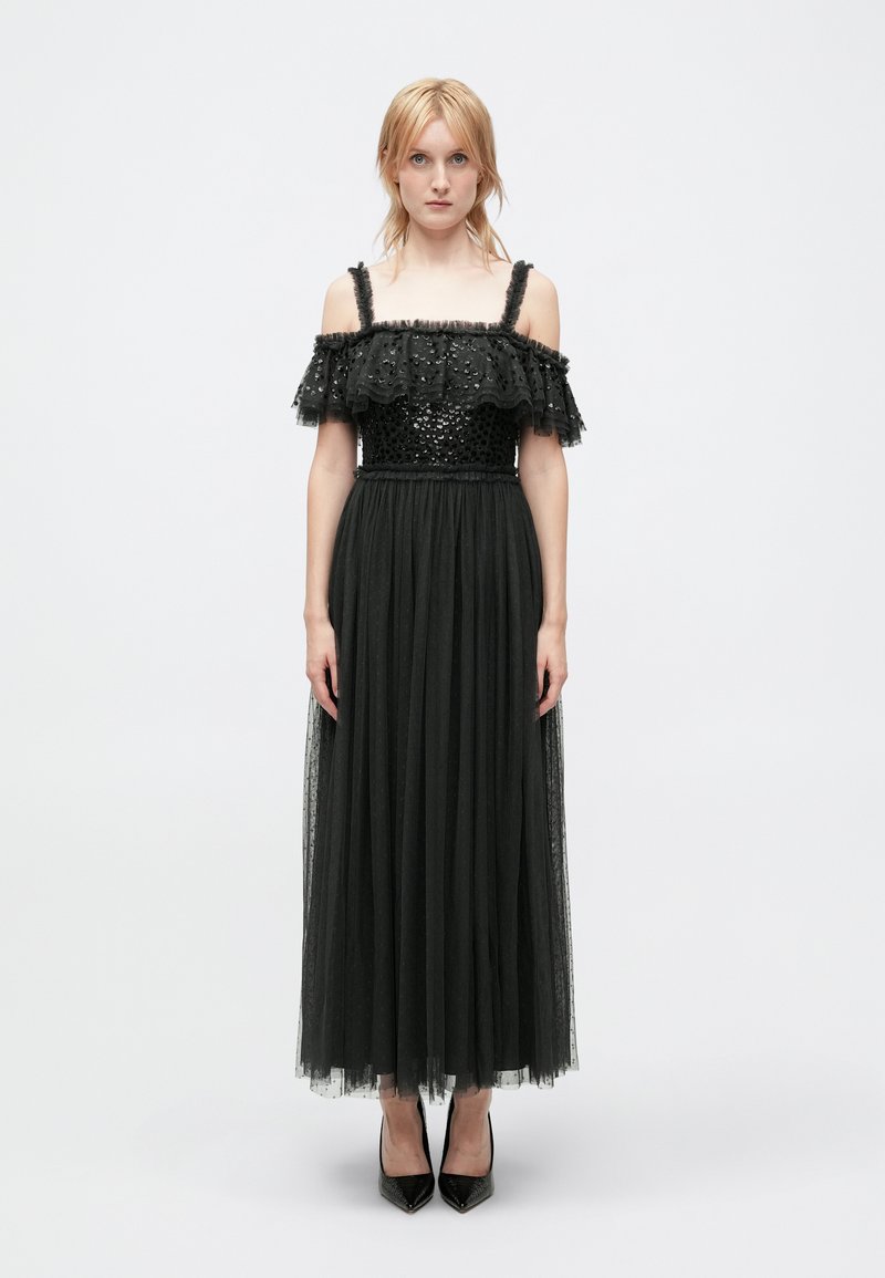 Robe noire avec des manches à volants dénudées, un corsage en sequins et une longue jupe en tulle. Associée à des talons aiguilles noirs.