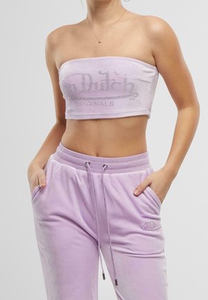 Kobieta ubrana w liliowy, bezramienny crop top Von Dutch oraz dopasowane, miękkie spodnie dresowe, z rękami w kieszeniach, na jednolitym szarym tle.