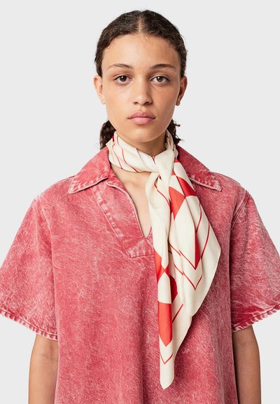 Jonge vrouw in een rood gewassen T-shirt met korte mouwen en een witte sjaal met rode geometrische patronen om haar hals geknoopt.