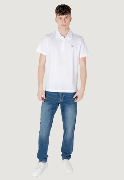 Chemise polo en coton blanc à col et à manches courtes, associée à un jean en denim bleu et des baskets blanches. Détail de logo subtil sur la chemise.