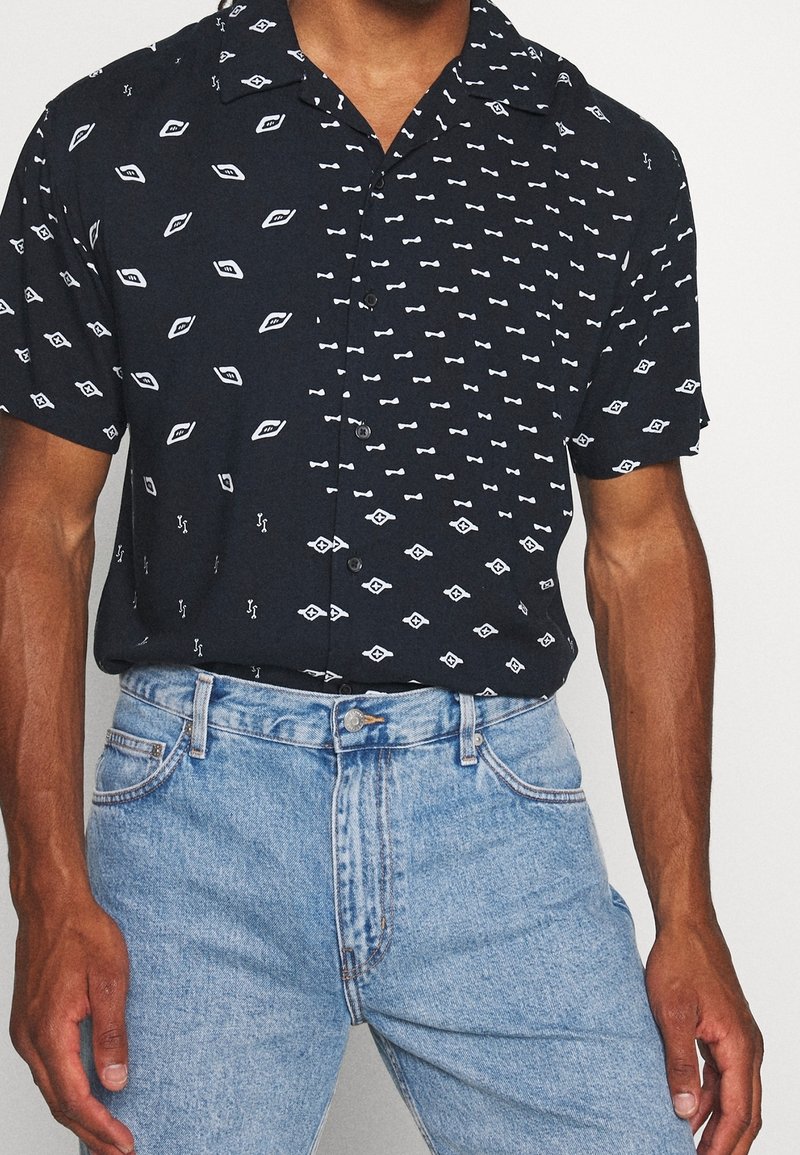 Kortärmad skjorta i svart med vita geometriska mönster. Mjuk, lätt tyg; kombinerad med högmidjade blå denimjeans.