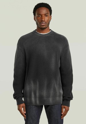 Uomo con capelli corti e ricci che indossa un maglione a costine grigio scuro e jeans scuri, in piedi contro uno sfondo neutro chiaro.