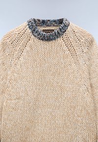 Kremowy sweter dzianinowy z teksturowanymi ściegami i niebieską kolorową żeberkowaną kołnierzykiem, ułożony na jasnym tle.
