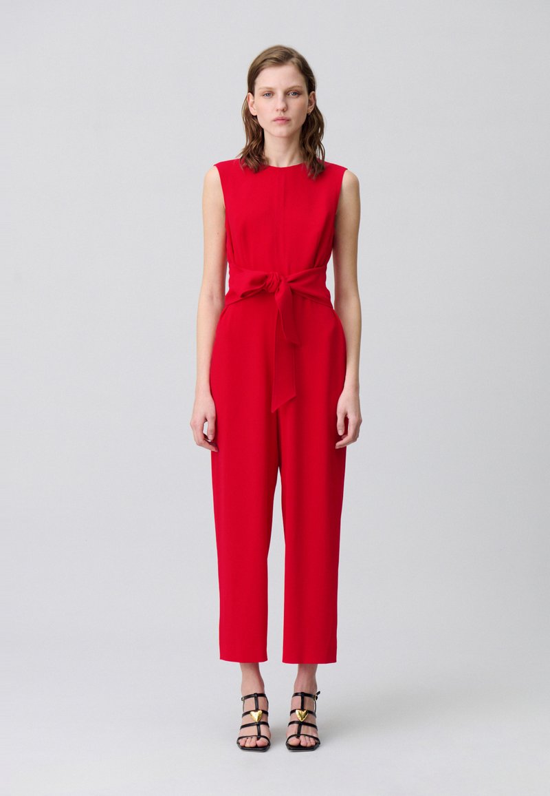Mono rojo sin mangas con un cinturón atado en la cintura, piernas rectas y una tela suave. Combinado con tacones negros con tiras y acentos dorados.