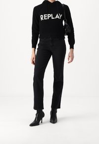 Schwarzer cropped Hoodie mit "REPLAY"-Schriftzug, kombiniert mit hochgeschnittenen schwarzen Jeans und spitzen schwarzen Stiefeletten. Glatte Textur insgesamt.