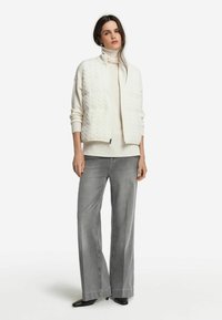 Kvinna klädd i vit kabelstickad zip-upp väst över krämfärgad polotröja, med grå vida jeans och svarta spetsiga skor, står upp.