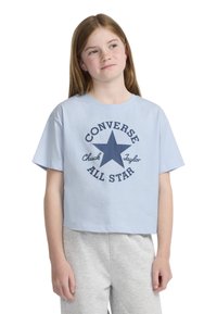 Trumpa šviesiai mėlyna medvilninė marškinėlių, kuriuose pavaizduota tamsiai mėlyna žvaigždė ir „Converse All Star“ tekstas apvalioje formoje. Pora su pilkais šortais.