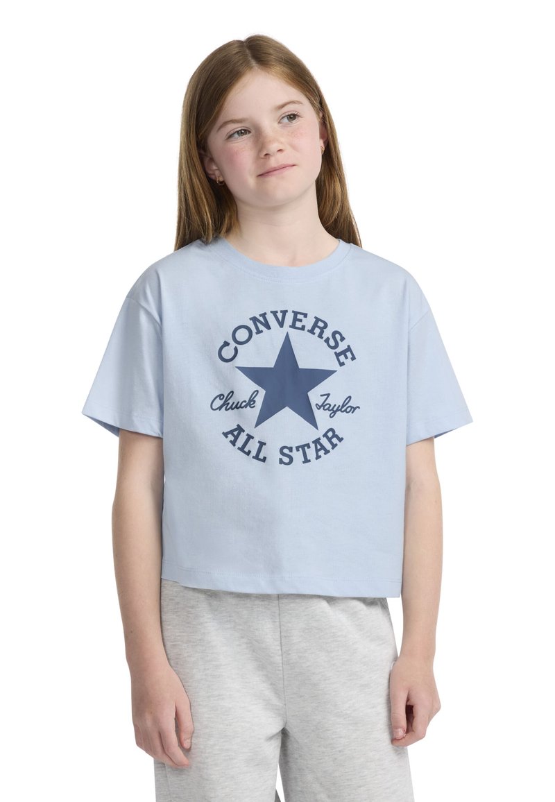 Trumpa šviesiai mėlyna medvilninė marškinėlių, kuriuose pavaizduota tamsiai mėlyna žvaigždė ir „Converse All Star“ tekstas apvalioje formoje. Pora su pilkais šortais.