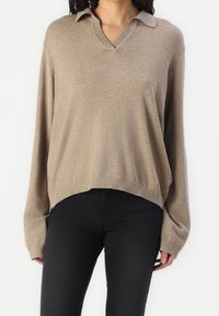 Pull-over en tricot beige avec un col en V et un col, doté de manches longues et d'une coupe décontractée, associé à un jean noir.