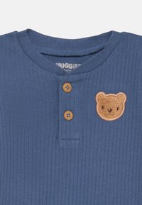 Top blu ribbed con scollatura rotonda, dotato di tre bottoni beige e una toppa con il volto di un orso marrone sul lato sinistro del petto.