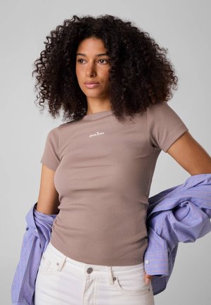 Jeune femme aux cheveux bouclés portant un t-shirt beige ajusté et un pantalon blanc, tenant une veste rayée bleue sur ses bras.
