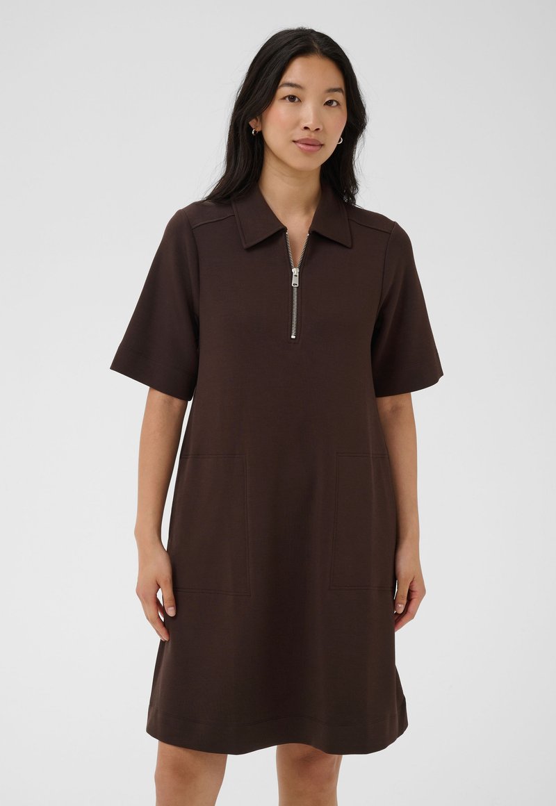 Femme portant une robe marron foncé jusqu'aux genoux, avec des manches courtes, un col, une fermeture éclair à l'avant et deux poches avant sur un fond clair uni.