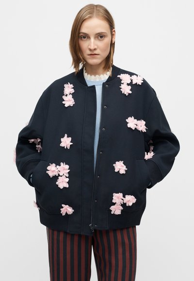 Jachetă bomber neagră decorată cu aplicații de flori roz, prevăzută cu manșete striate, închidere cu fermoar frontal și buzunare laterale. Se văd pantaloni cu dungi.