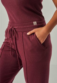 Nahaufnahme einer Person, die gerippte, burgunderrote Strickhosen mit Kordelzugbund trägt und deren Hand in der Seitentasche steckt.