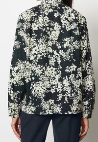 Blouse fleurie noire avec motifs floraux blancs. Dotée d'un col, de manches longues et d'une coupe décontractée. Texture de tissu lisse.
