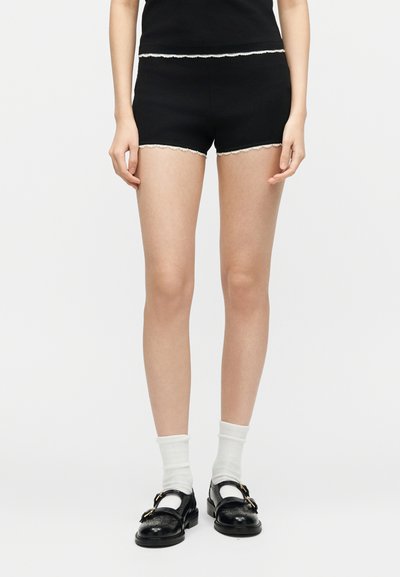 Personne portant un short noir avec un bord en dentelle blanche, des chaussures Mary Jane noires et des chaussettes blanches, debout devant un fond blanc uni.