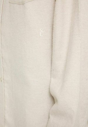 Beige skjorte med knapper lavet af blødt stof, med et subtilt tekstureret mønster og et broderet logo på lommen.