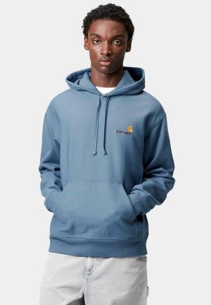 Sudadera azul hecha de tela suave, con un bolsillo frontal, capucha con cordón y un pequeño logotipo bordado en naranja. Combinada con jeans gris claro.