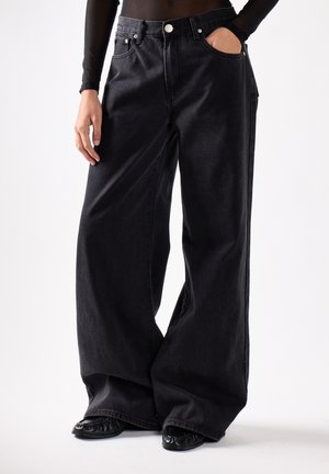 Person iført oversized sorte vidbenede jeans og sorte sko med den ene hånd i lommen og den anden afslappet ved siden.
