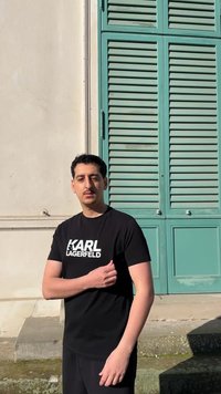 Mann i svart Karl Lagerfeld T-skjorte står ute ved siden av en beige vegg og et teal-farget vindu med skodder i sollys.