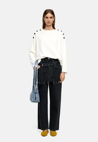 Jasno-beżowy, oversize sweter z czarnymi guzikami na ramionach, zestawiony z czarnymi, wysokimi spodniami z szerokimi nogawkami, niebieską torbą na ramię i żółtymi butami.
