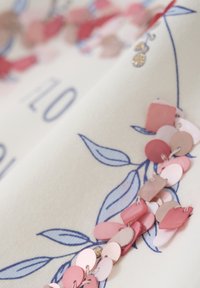 Broderad tyg med ljus creamfärgad bas, prydd med blå bladmönster och rosa paljetter i olika former sydda längs kanten.
