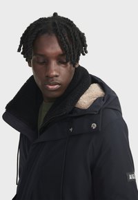 Veste bleu marine avec un col en polaire noir, dotée de boutons-pression en métal et d'un patch logo sur la manche. Tissu en mélange de coton et de polyester.