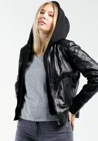 Schwarze Lederjacke mit Kapuze, glatte Textur, Reißverschluss und Seitentaschen. Über einem grauen V-Ausschnitt-Pullover und dunkelblauen Jeans getragen.