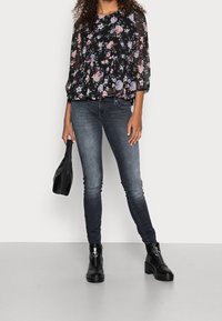 Schwarze Bluse mit floralem Muster und transparenten langen Ärmeln, kombiniert mit dunklen Skinny-Jeans. Dazu eine kleine schwarze Handtasche und knöchelhohe schwarze Stiefel.