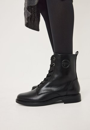 Svarta läderankelboots med snörning, rund tå och gummisula. Har en cirkulär logotyp präglad på sidan.