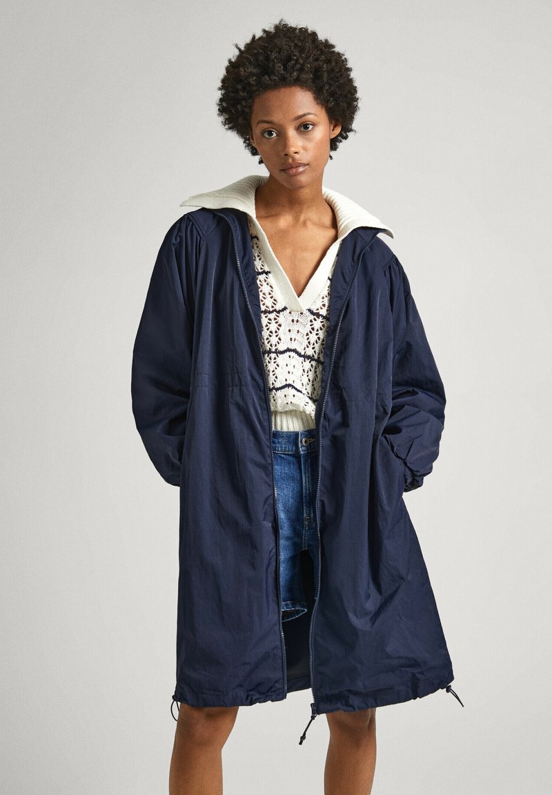 Veste légère bleu marine avec un col texturé blanc, dotée d'une fermeture éclair à l'avant et d'un ourlet à cordon de serrage. Portée sur un pull crème et un short en denim.