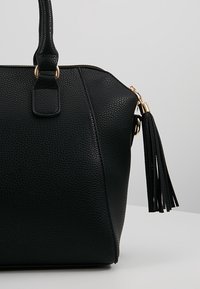 Sac à main noir texturé avec une poignée supérieure, des éléments en métal doré, une fermeture éclair et un pompon noir attaché au curseur de la fermeture.