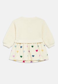 Φόρεμα εκρού με αφράτο, υφασμάτινο μπούστο και φαρδιά φούστα με σχέδιο πολύχρωμες καρδιές σε ανοιχτό φόντο.