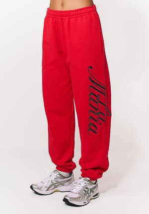 Pantaloni sport roșii cu talie elastică, având un print negru scris în cursiv pe piciorul stâng. Materialul este neted și texturat.