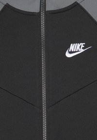 Giacca nera con zip e dettagli grigi, dotata di una zip argentata e un logo Nike ricamato in bianco sul petto sinistro. Materiale liscio e strutturato.