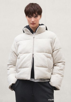 Jeune personne aux cheveux courts et foncés portant une veste matelassée beige clair par-dessus un sweat à capuche sombre, mains dans les poches, sur un fond clair uni.
