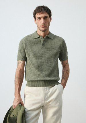 Hombre con tatuajes lleva un polo de manga corta verde oliva texturizado y pantalones crema, sosteniendo una chaqueta verde oliva a juego en la mano izquierda.