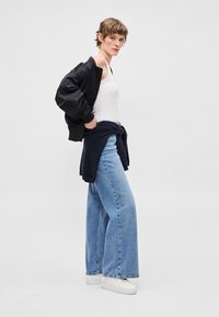 Μαύρο μπουφάν bomber, λευκό αμάνικο τοπ, ανοιχτό μπλε τζιν με φαρδιά μπατζάκια και λευκά αθλητικά παπούτσια. Το μοντέλο στέκεται πλάγια με μπλε σκούρο πουλόβερ δεμένο στη μέση.