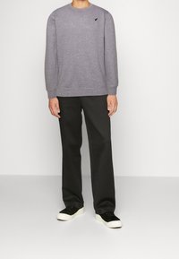 Felpa grigia con scollatura tonda e piccolo logo scuro sul lato sinistro del petto, abbinata a pantaloni neri e scarpe da ginnastica nere. Tessuto dalla texture liscia.