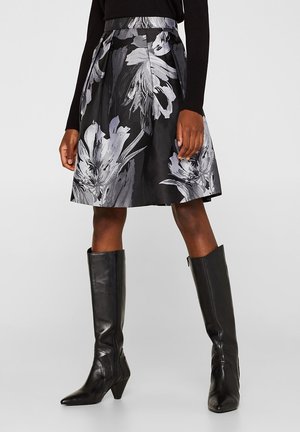 Frau trägt einen schwarz-grauen floralen A-Linien-Rock kombiniert mit schwarzen kniehohen Lederstiefeln und einem schwarzen Langarmshirt.