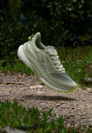 Pantof de alergare pe traseu Scarpa verde deschis, surprins în aer deasupra unui drum cu pietriș, înconjurat de iarbă și copaci, evidențiind aderența și elementele de design.