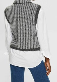 Gilet lavorato a maglia a strisce grigie e nere, con un colletto alto e una texture a costine, indossato sopra una camicia bianca. I bordi sono delicatamente frangiati.
