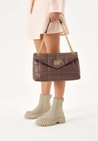 Sac à main matelassé marron avec une anse en chaîne dorée et un détail de logo, associé à des bottines en suède gris clair à bout arrondi avec une semelle épaisse.
