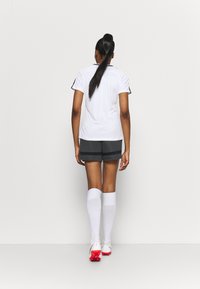 Bílé sportovní tričko s černými akcenty, šedé šortky, bílé podkolenky a červené kopačky, pohled zezadu. Hladká tkanina, sportovní design.