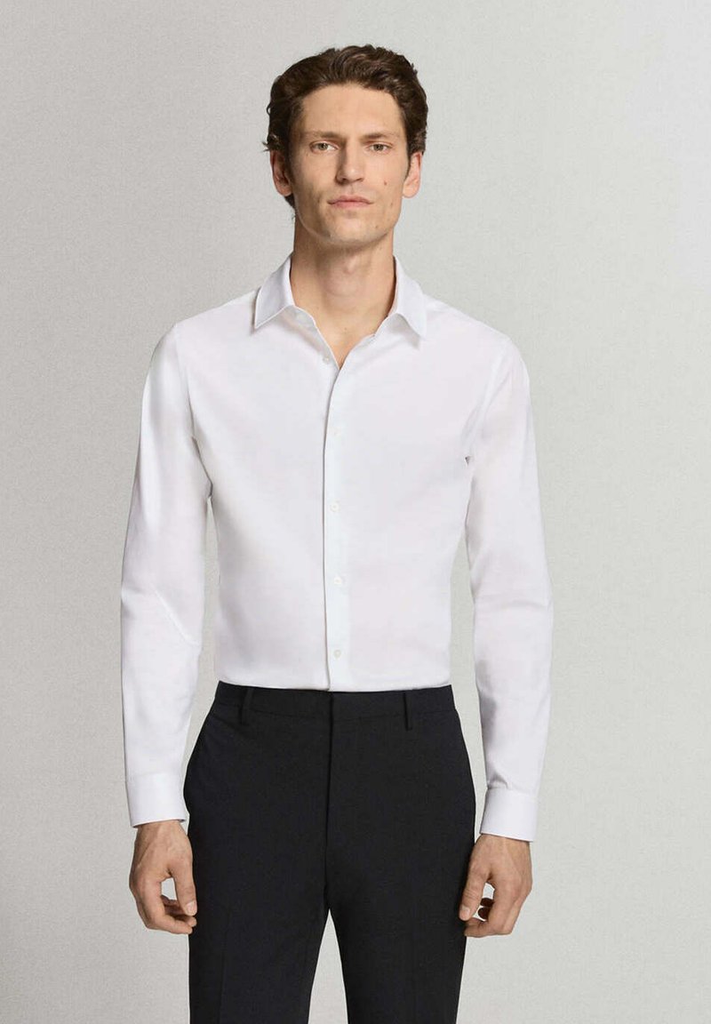 Homme portant une chemise blanche à manches longues et un pantalon noir, debout devant un fond gris clair uni.