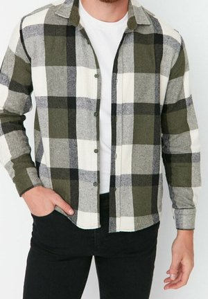Homme portant une chemise à carreaux verte, noire et blanche boutonnée sur un t-shirt blanc avec un pantalon noir, main dans la poche.