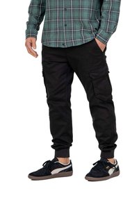 Pantalones cargo negros con bolsillos laterales y puños elásticos, combinados con una camisa de cuadros verde y unos zapatos deportivos de ante oscuros.