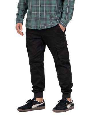 Black cargo pants with side pockets and elastic cuffs, paired with a green plaid button-up shirt and dark suede sneakers.