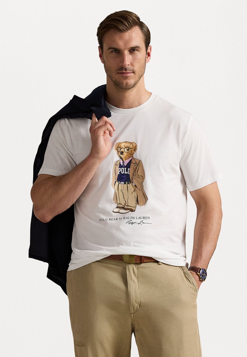 Bela bombažna majica z grafiko medveda v blazerju in polo majici. Model drži mornarsko jakno čez ramo, oblečen je v kaki hlače.